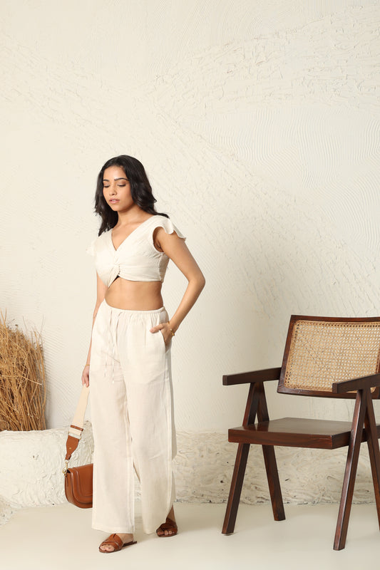 Woman in a white outfit standing in a minimalistic room with a wooden chair and textured wall.