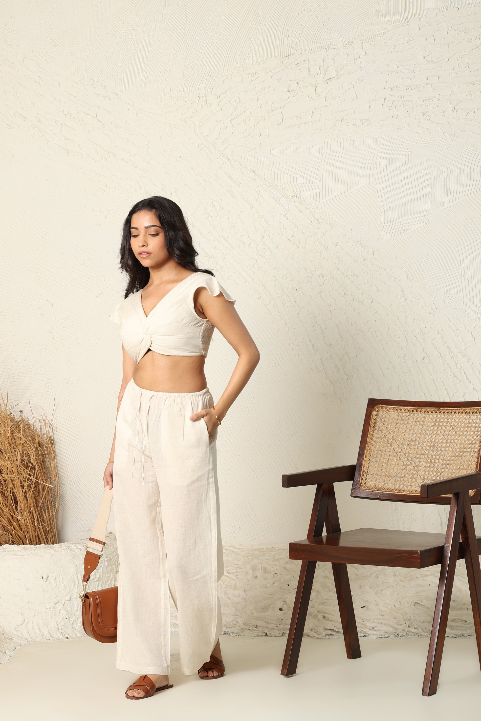 Woman in a white outfit standing in a minimalistic room with a wooden chair and textured wall.