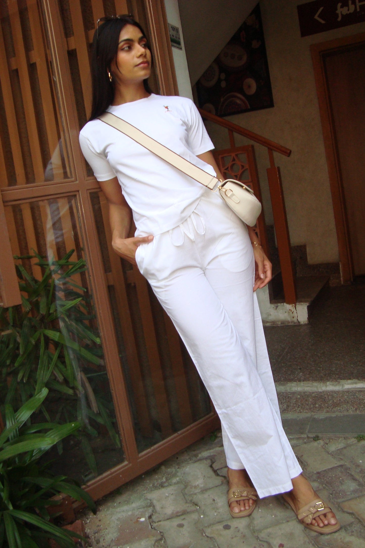 Woman in white outfit with beige bag standing outdoors near a building entrance.